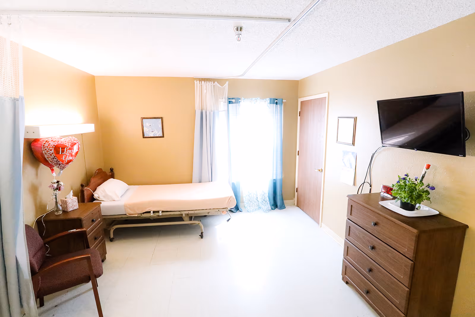 A bright resident bedroom with a single hospital-style bed, dresser, wall-mounted TV, chair, and a window with curtains.
