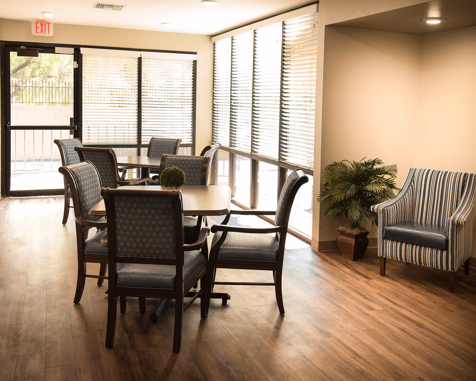 Sunlit common dining area with multiple tables and chairs, large windows, and an upholstered armchair beside a potted plant.