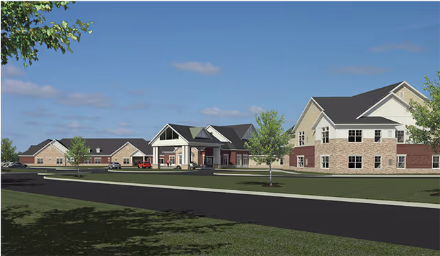 Front exterior of a low-rise senior living facility with multiple connected brick-and-siding buildings, a covered porte-cochere entrance, driveway and landscaped lawn under a blue sky.