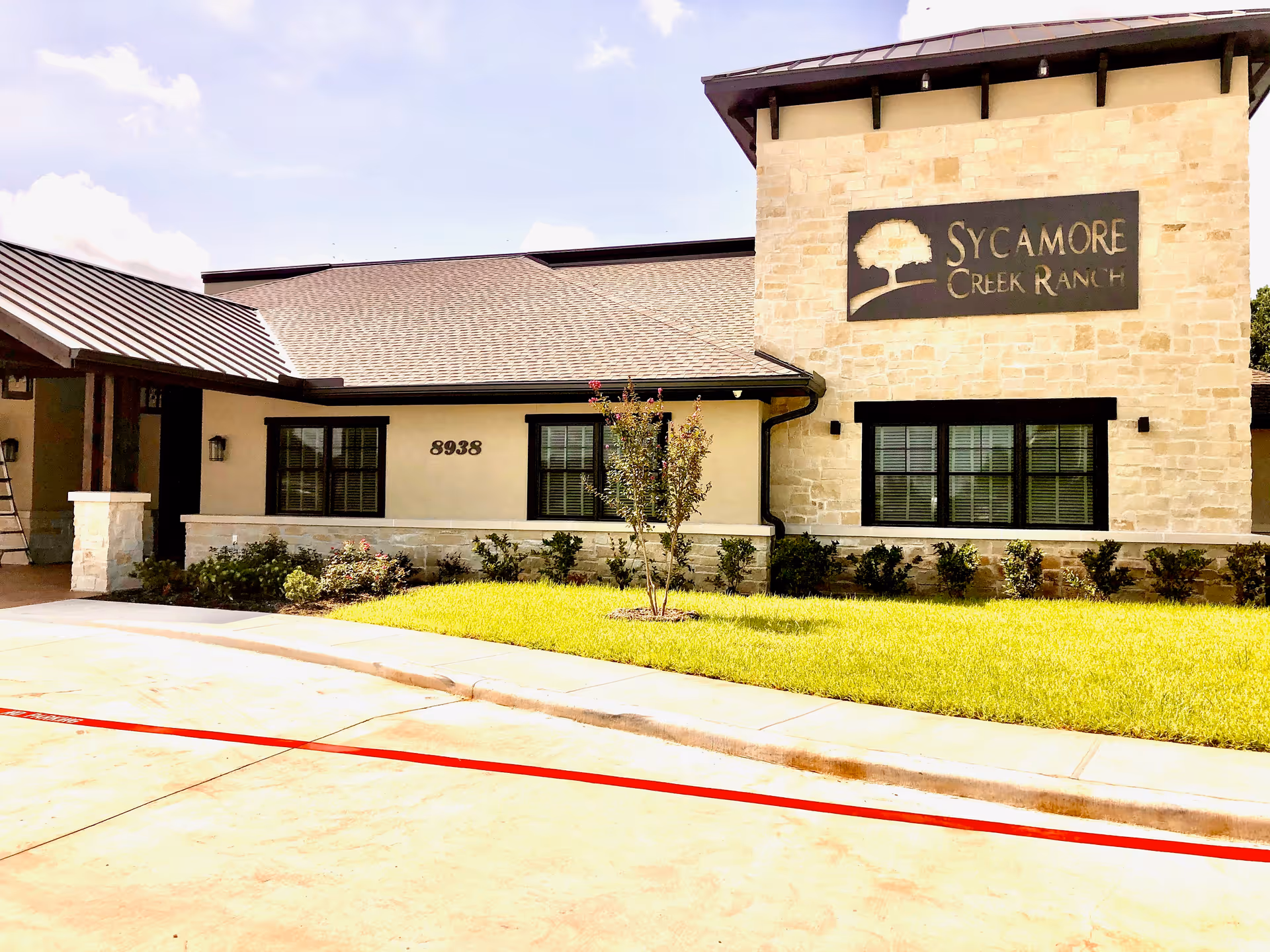 Exterior view of Sycamore Creek Ranch building with stone and beige walls, black framed windows, a small tree and bushes in front, and a driveway with a red painted curb.