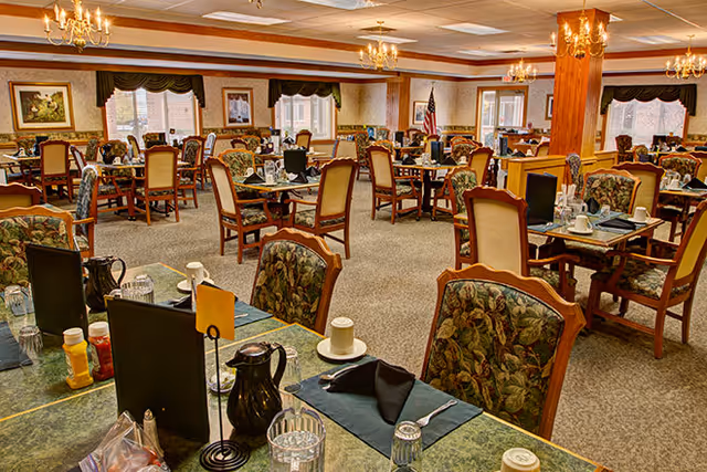 A spacious dining room with multiple tables and chairs arranged neatly. The chairs have floral upholstery and wooden frames. Each table is set with placemats, black napkins, cups, glasses, and condiments. The room has chandeliers hanging from the ceiling, large windows with valances, and framed artwork on the walls. An American flag is visible in the background near a window.