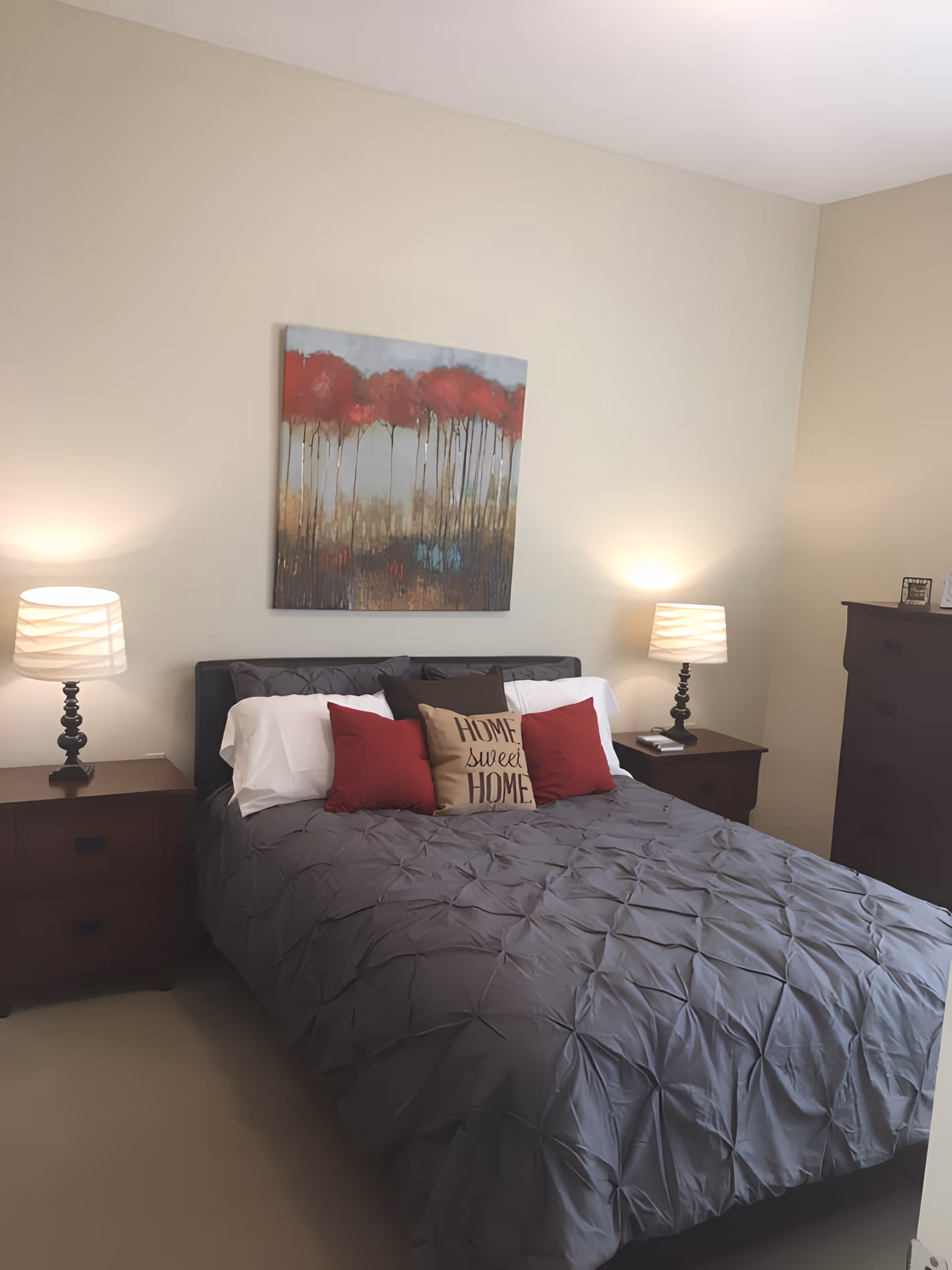 A bedroom with a neatly made bed featuring a gray comforter and several pillows, including one with the text 'HOME sweet HOME'. There are two wooden nightstands on either side of the bed, each with a lamp. Above the bed hangs a painting of red trees with a muted background. A wooden dresser is visible on the right side of the room.