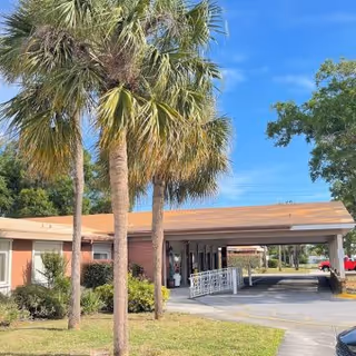 Exterior view of a single-story building with a covered entrance and a driveway. There are palm trees and other greenery in the foreground under a clear blue sky.