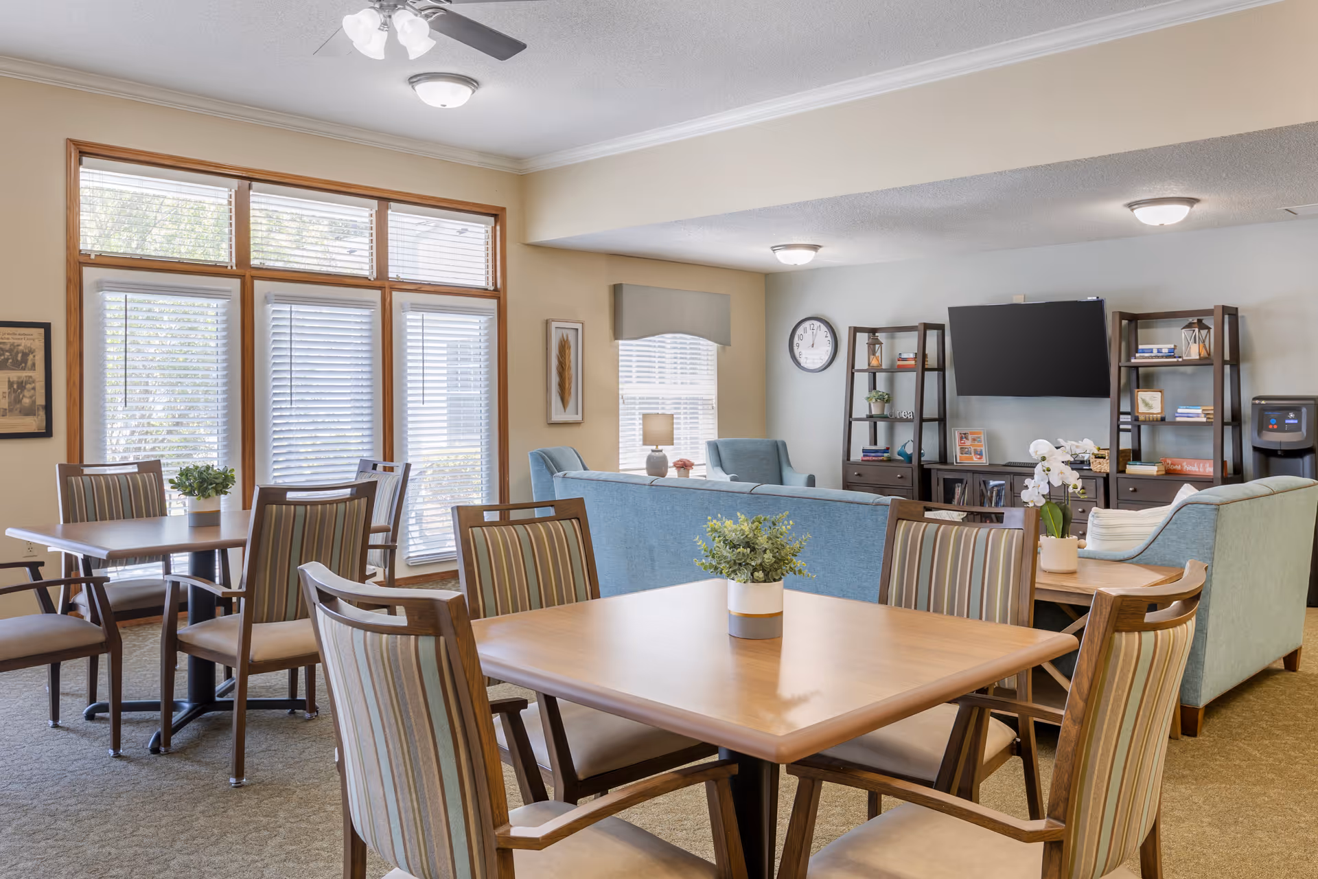 Bright communal dining and lounge area with wooden tables and chairs, blue sofas, a wall-mounted TV, and large windows.