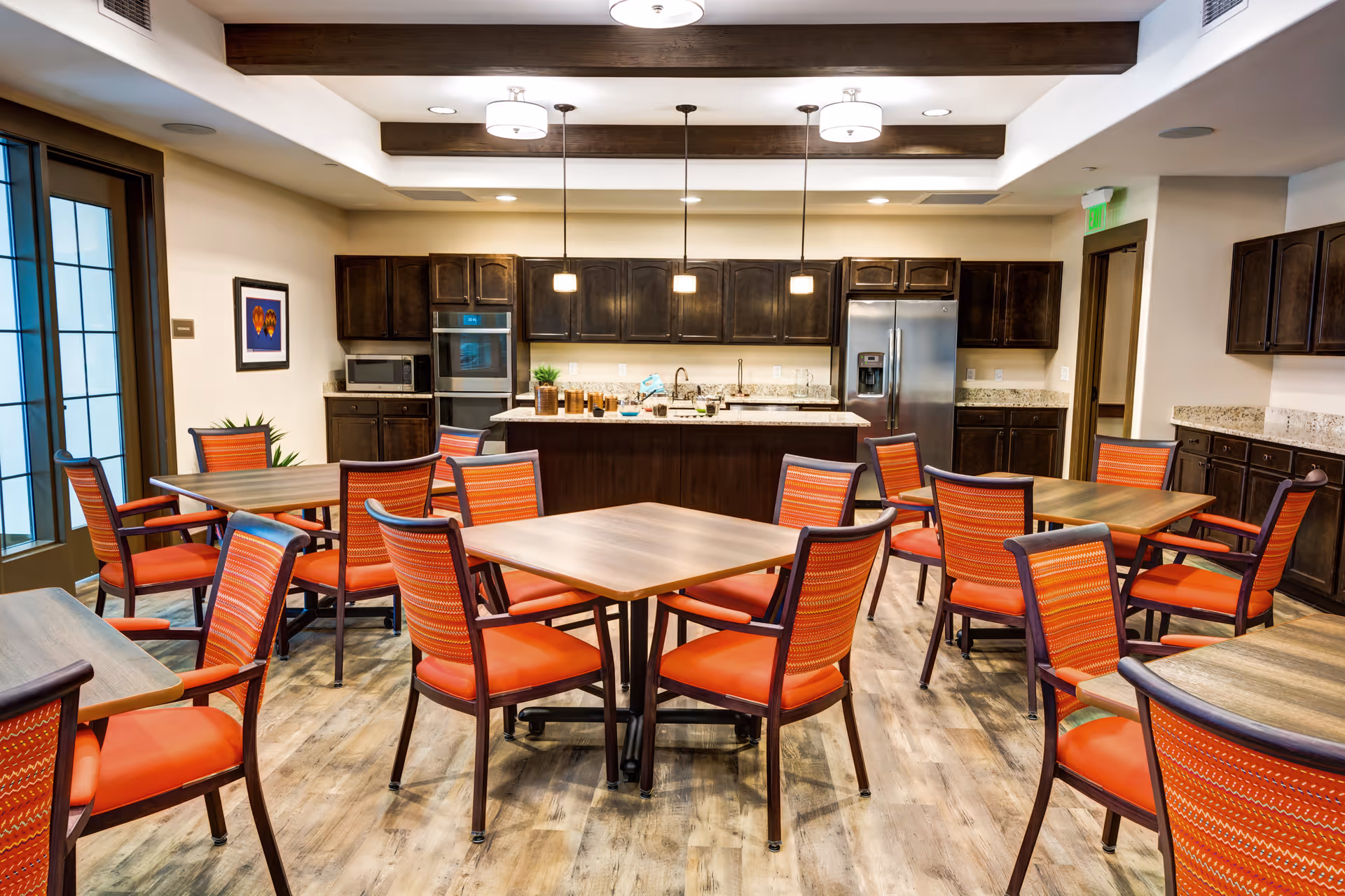 A bright and spacious dining area in a senior living facility featuring multiple wooden tables surrounded by orange cushioned chairs. The room has a modern kitchen in the background with dark wood cabinets, stainless steel refrigerator, microwave, and oven. Pendant lights hang from the ceiling above a central island, and the floor is covered with light wood-style flooring.