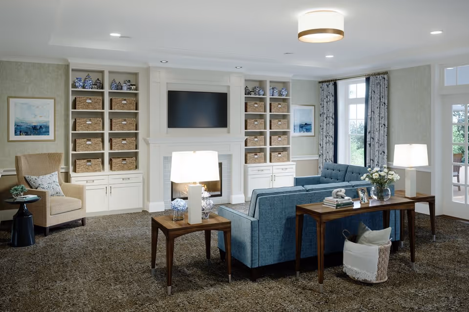 A cozy living room with a blue sofa, two wooden side tables with lamps, a beige armchair with a pillow, built-in white shelves filled with wicker baskets and decorative blue and white vases, a fireplace with a mounted flat-screen TV above it, framed artwork on the walls, large windows with patterned curtains, and a carpeted floor.