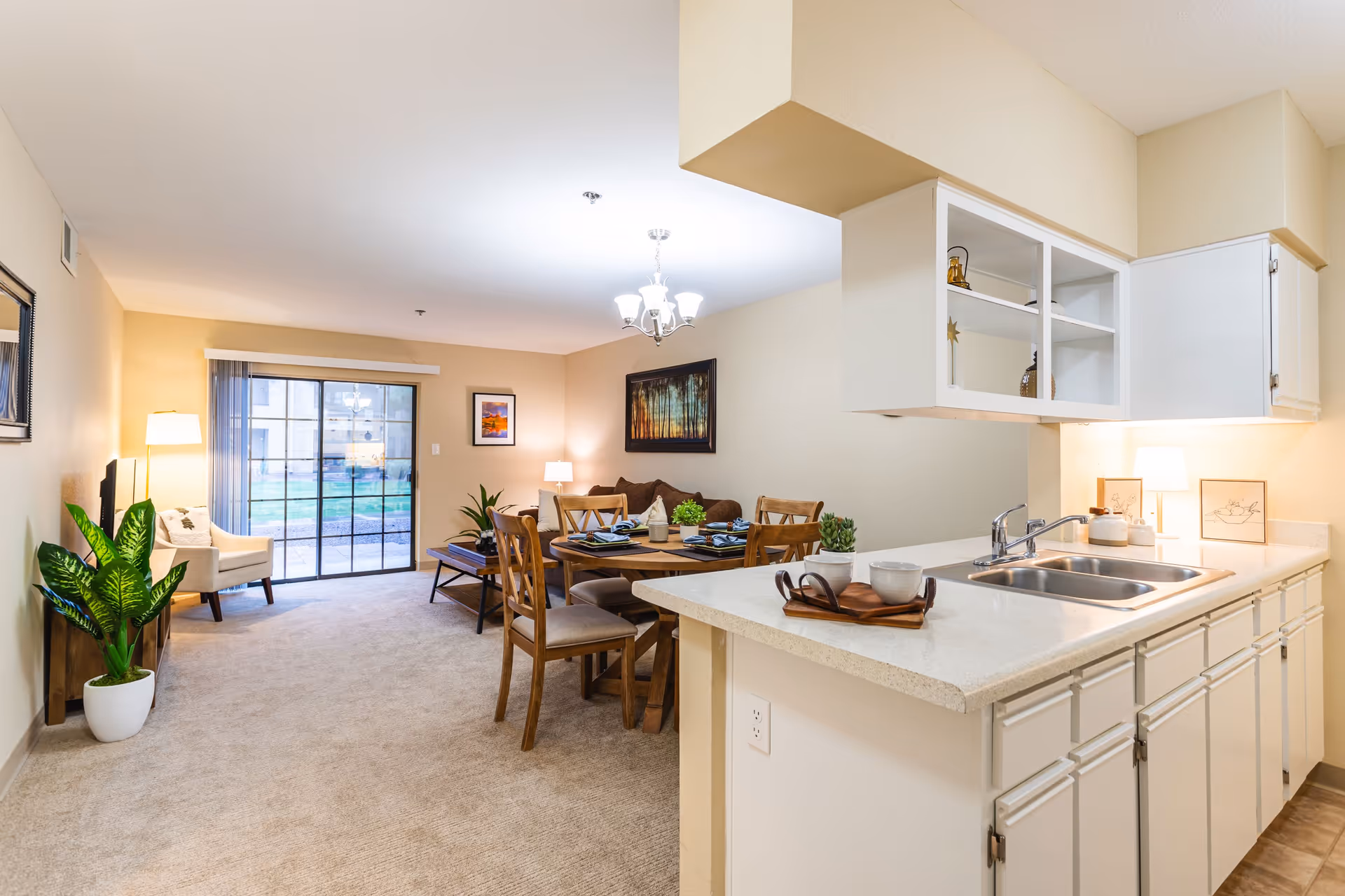Bright and spacious interior of a senior living facility apartment showing a kitchen with white cabinets and a double sink in the foreground. Beyond the kitchen is a dining area with a wooden table set for four, a living area with a brown sofa, a coffee table, a white armchair, and a large sliding glass door leading outside. The room is decorated with plants, framed pictures, and warm lighting.