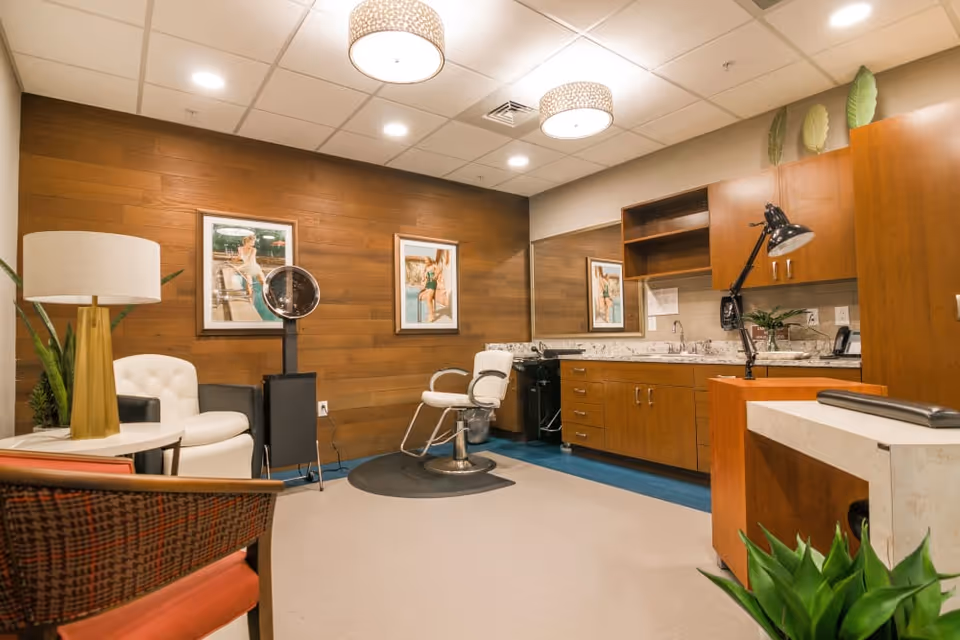 Interior room with a salon chair, a white cushioned chair, a lamp, wooden cabinets with a countertop and sink, a large mirror, framed pictures on a wooden paneled wall, and some plants.