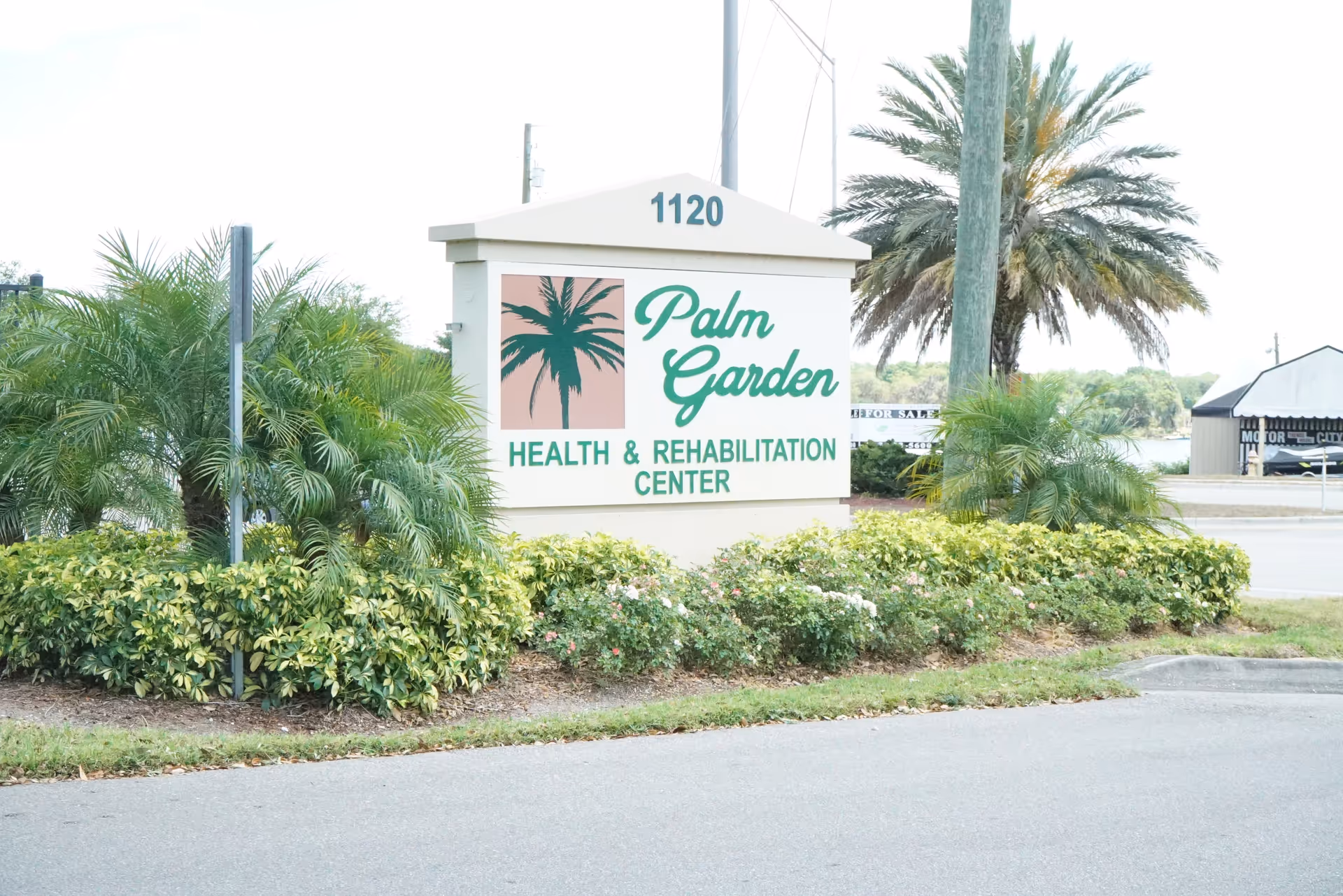 Entrance sign reading 'Palm Garden Health & Rehabilitation Center' surrounded by palm trees and landscaping.