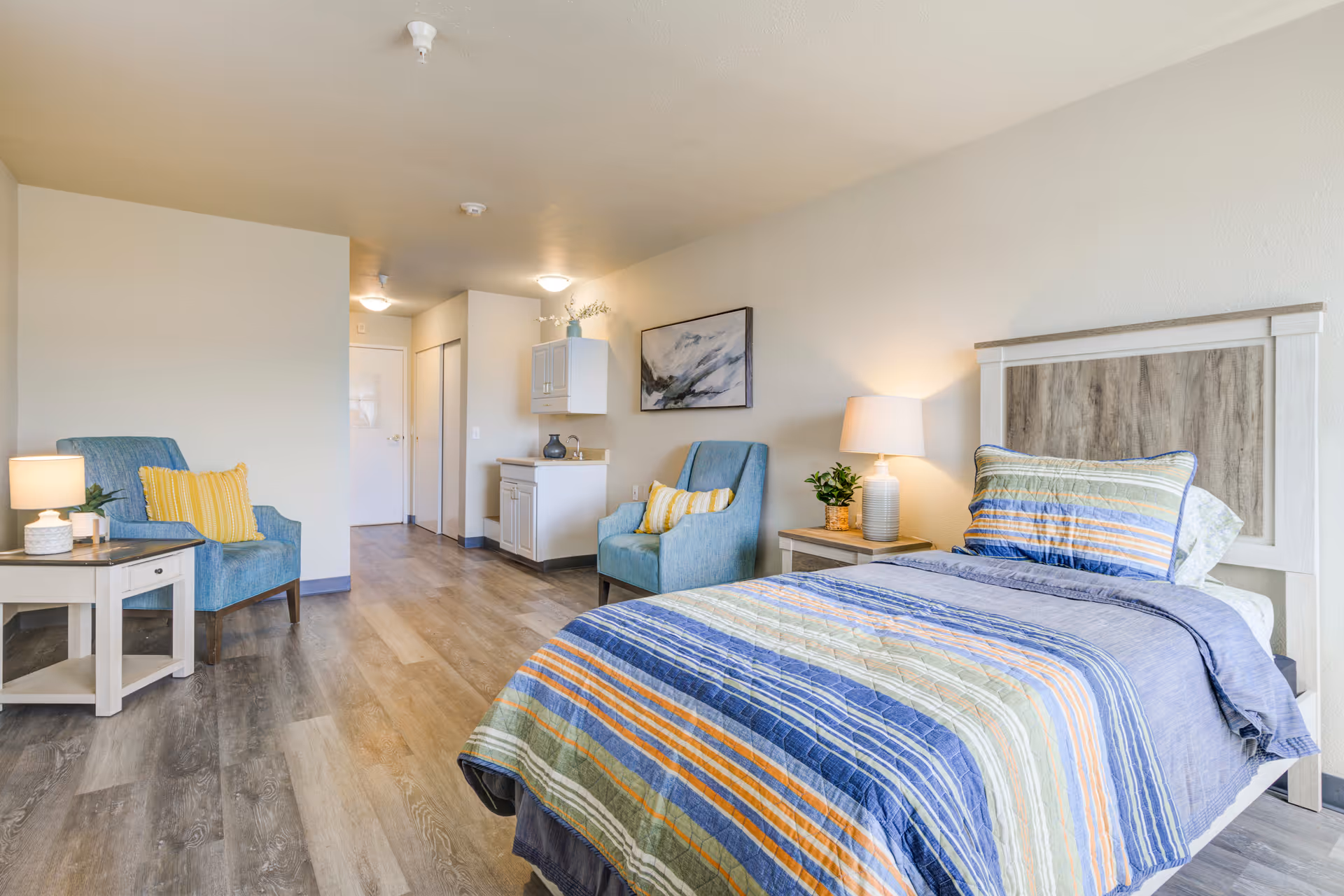 A bright furnished bedroom studio with a bed, two armchairs, nightstands, lamps, and a small kitchenette area.