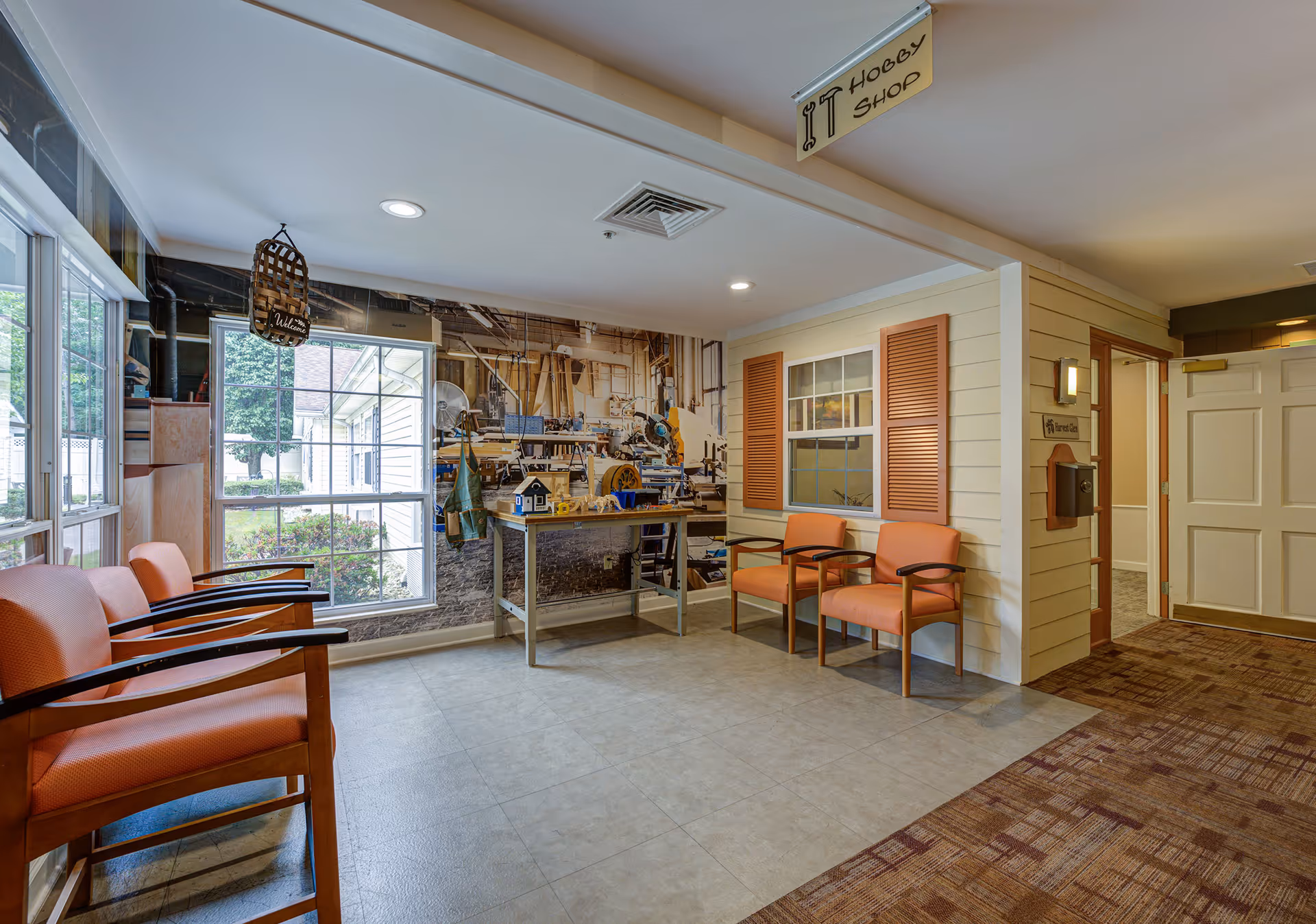A cozy indoor hobby shop area in a senior living facility with four orange cushioned chairs, a table with miniature woodworking models, a large wall mural depicting a workshop, and a sign overhead reading 'Hobby Shop'. There is a window with brown shutters and a door leading to another room.