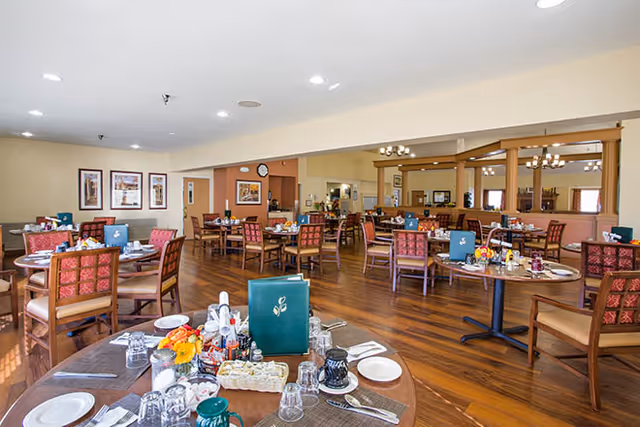 Spacious dining room with round tables set for meals, wooden chairs, and a serving area in the back.