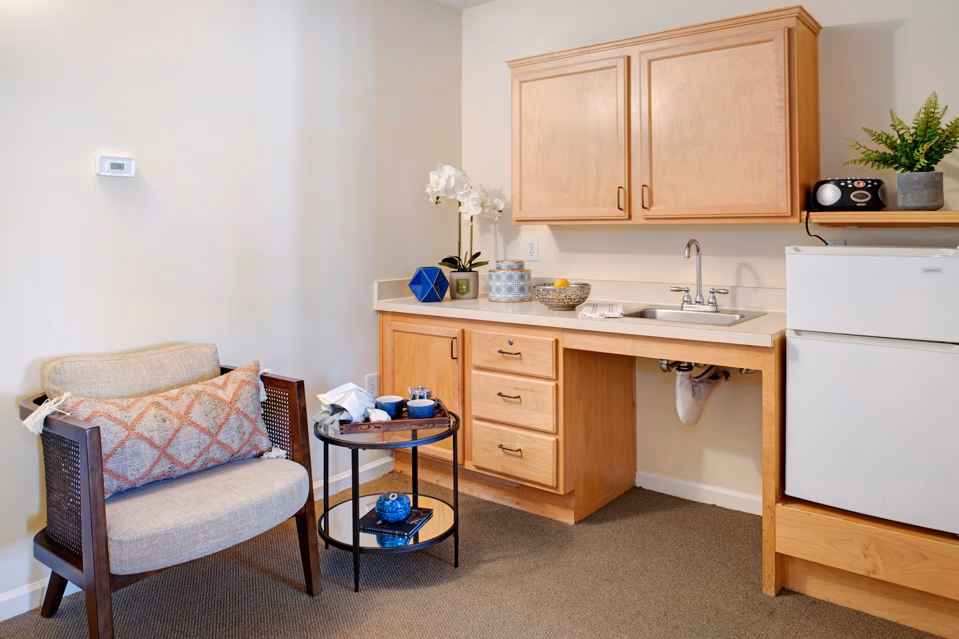 Studio-style kitchenette and seating area with light wood cabinets, a sink and mini-fridge beside an upholstered chair and small round table.