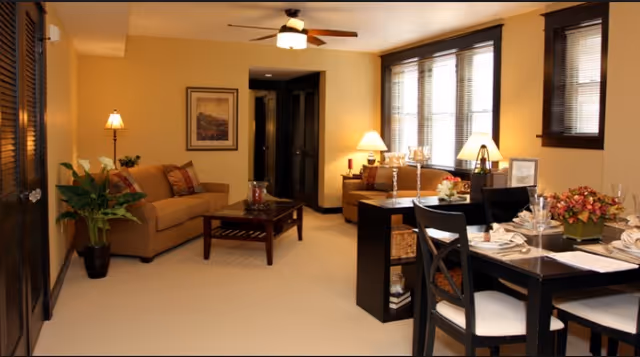 A warmly lit living and dining area with beige walls and carpet. The living room features a tan sofa with decorative pillows, a wooden coffee table, a floor lamp, and framed artwork on the wall. The dining area has a dark wooden table set with plates, glasses, and napkins, surrounded by chairs. Large windows with blinds allow natural light into the space, and ceiling fan with light is mounted above.