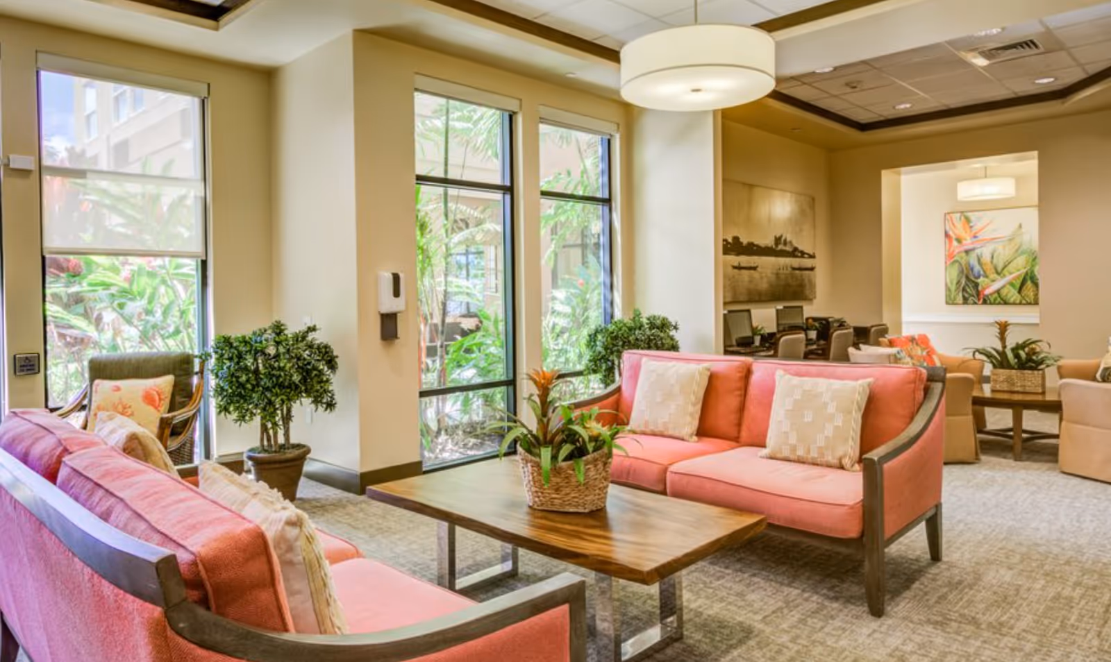 Bright communal lounge with pink sofas, wooden coffee tables, potted plants, and large windows overlooking greenery.