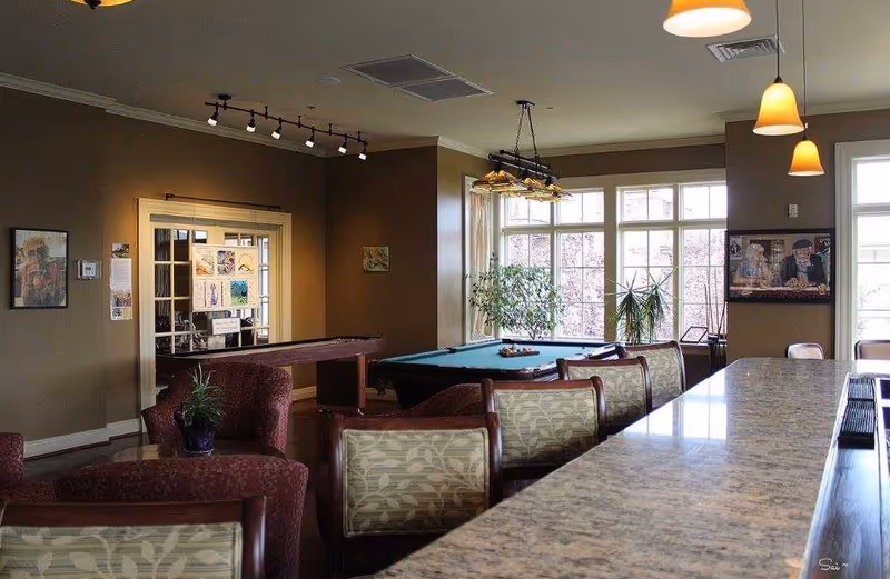 A cozy senior living common room with a pool table, plush seating, a long bar counter, pendant lights, and large windows with plants.