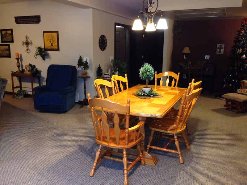 A cozy interior room with a wooden dining table and six matching chairs in the center. The table has a small green plant centerpiece. To the left, there is a blue armchair and various wall decorations including framed pictures and a decorative cross. In the background to the right, a Christmas tree is visible along with a lamp and other furniture. The room is carpeted and softly lit by a ceiling light fixture above the table.