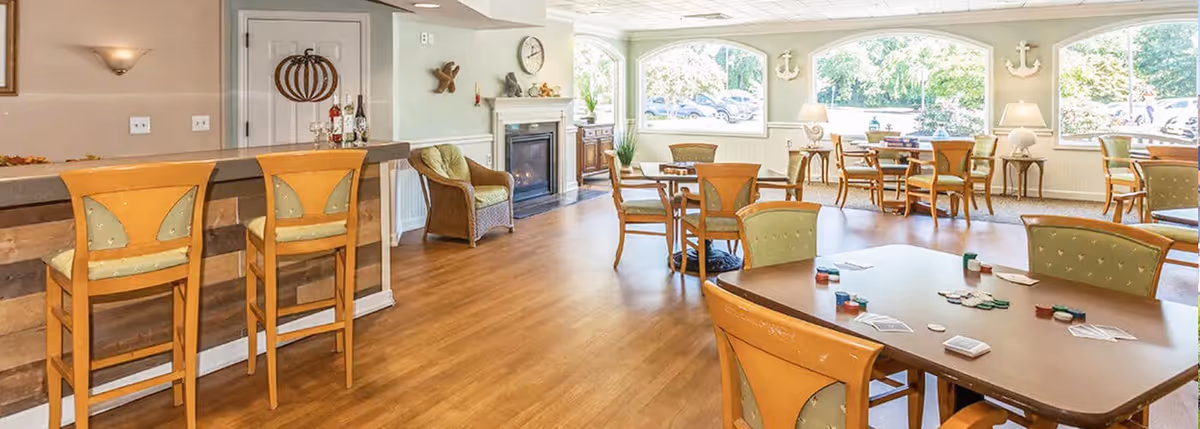 A spacious, well-lit common area in Milford Place featuring wooden floors, multiple tables and chairs arranged for social activities, a bar counter with two high chairs, a fireplace, and large windows letting in natural light with a view of parked cars outside.