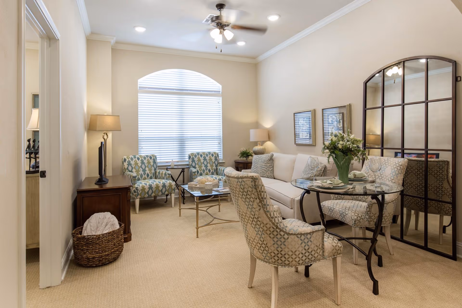 A cozy living room with beige walls and carpet, featuring a white sofa with patterned cushions, two patterned armchairs around a glass coffee table, and a small round glass dining table with two patterned chairs. A large decorative mirror with a black grid frame leans against the wall, and a window with white blinds lets in natural light. There are two table lamps on wooden side tables and a ceiling fan with lights above.
