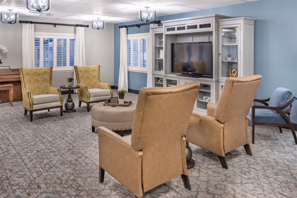Bright communal living room with multiple armchairs around an ottoman, a large TV in a white entertainment center, and windows with curtains.