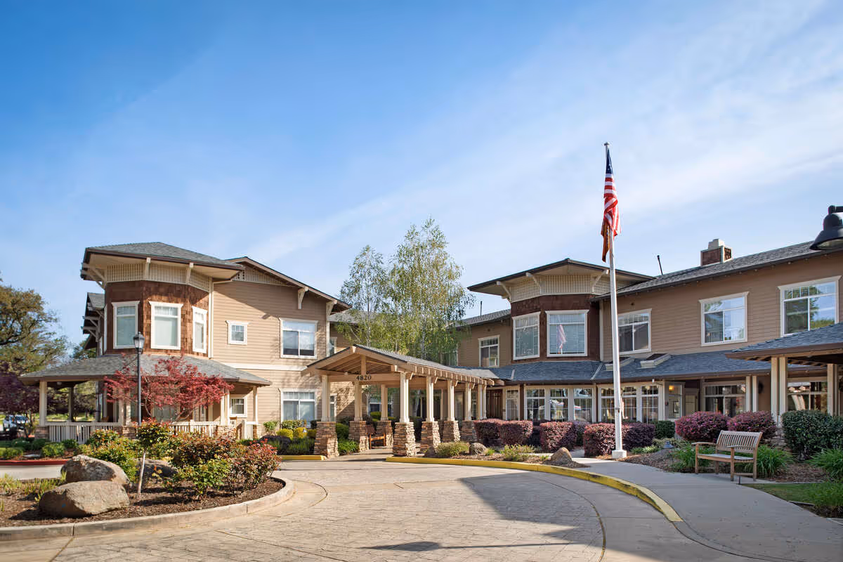 Exterior view of a senior living facility building with beige siding and stone accents, a covered entrance with stone pillars, landscaped bushes and trees, an American flag on a flagpole, and a clear blue sky.