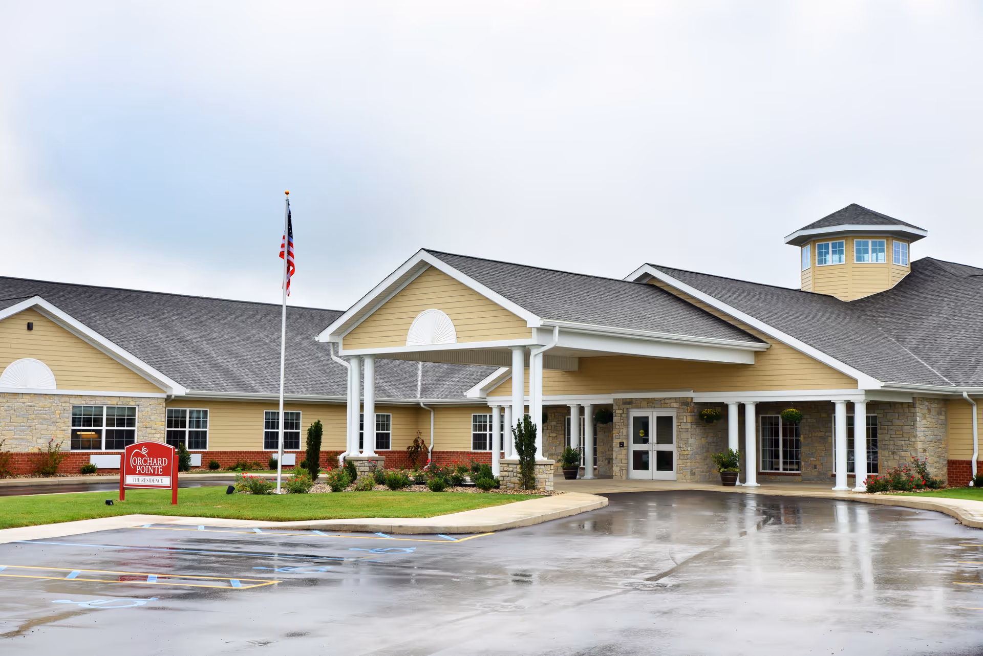 Front exterior view of Orchard Pointe Health Campus building with a covered entrance, stone and yellow siding, an American flag on a flagpole, and a wet parking lot in front.