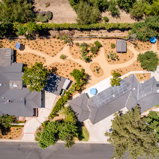 Aerial view of a senior living facility showing buildings with gray roofs, pathways winding through landscaped gardens with trees and shrubs, and outdoor seating areas with umbrellas.