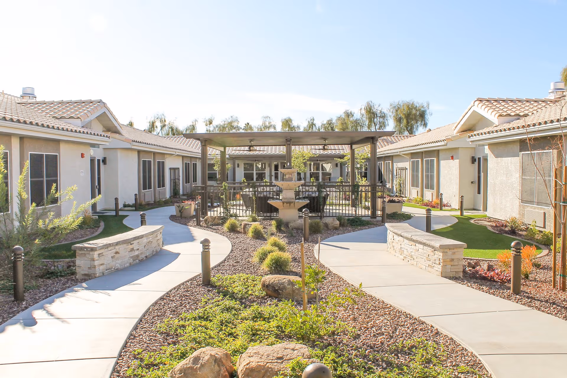 Outdoor courtyard area at Fairmont Village featuring a paved walkway that curves around landscaped garden beds with rocks and greenery. There is a central covered seating area with a multi-tiered stone fountain, surrounded by single-story buildings with tiled roofs and multiple windows.