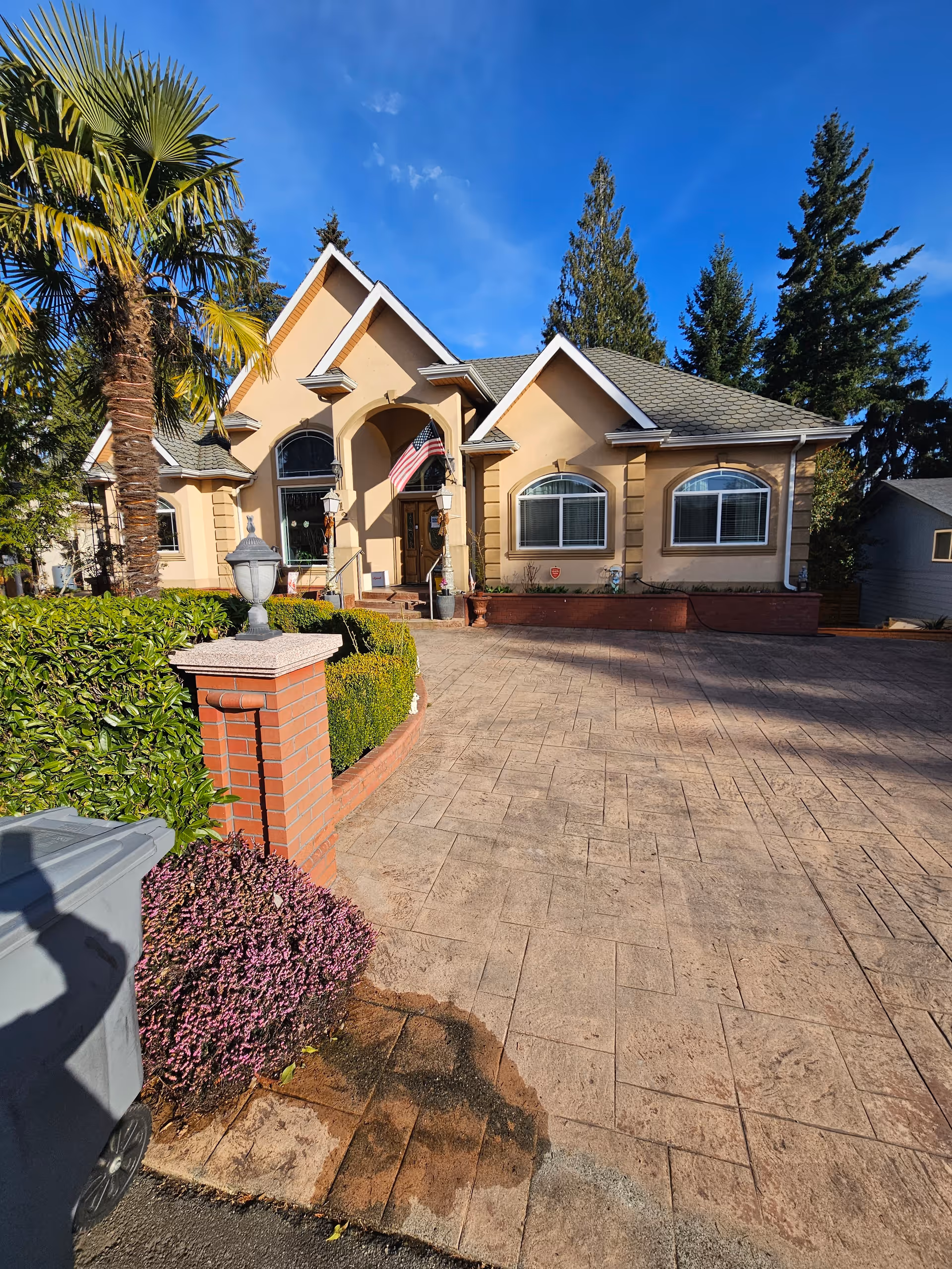 Front exterior view of a beige-colored residential building with a gabled roof, large windows, and an American flag hanging near the entrance. The driveway is paved with stamped concrete, and there are landscaped bushes, a palm tree, and other greenery around the property under a clear blue sky.