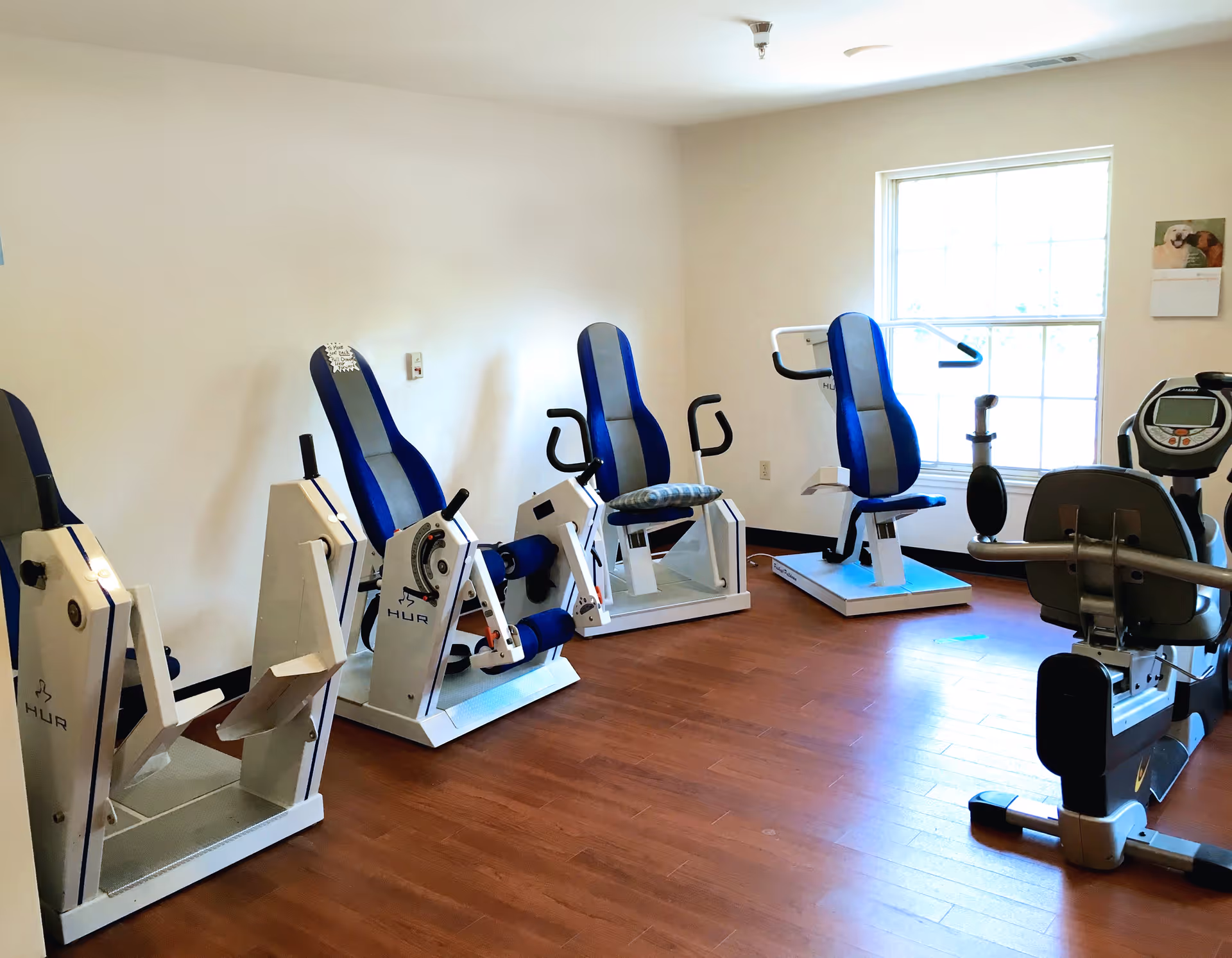 A small fitness room with several exercise machines featuring blue and gray padded seats. The room has wooden flooring, white walls, and a large window letting in natural light.