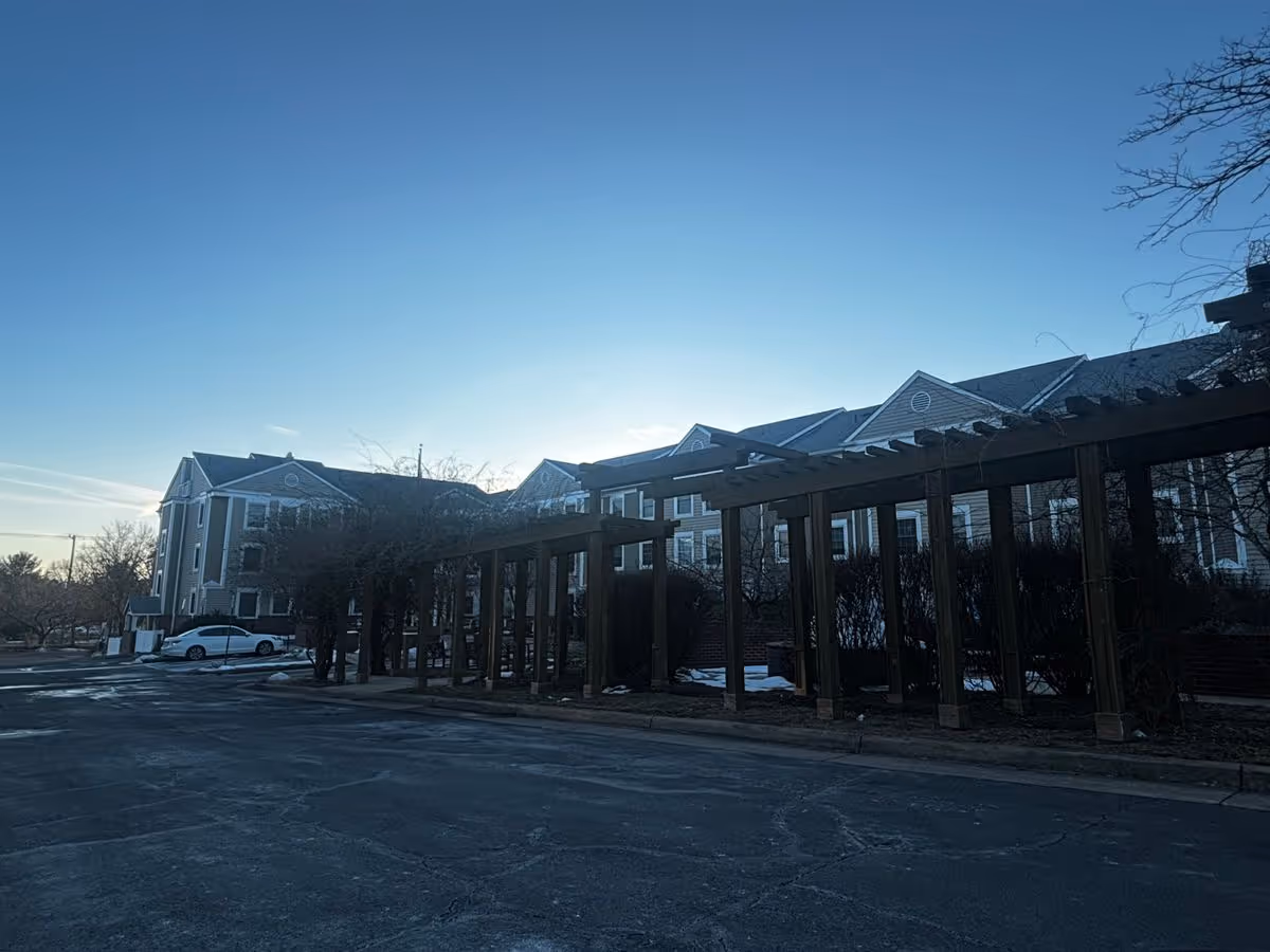 Exterior of a multi-story senior living building with a wooden pergola, parking area, and a car under a clear sky at dawn or dusk.