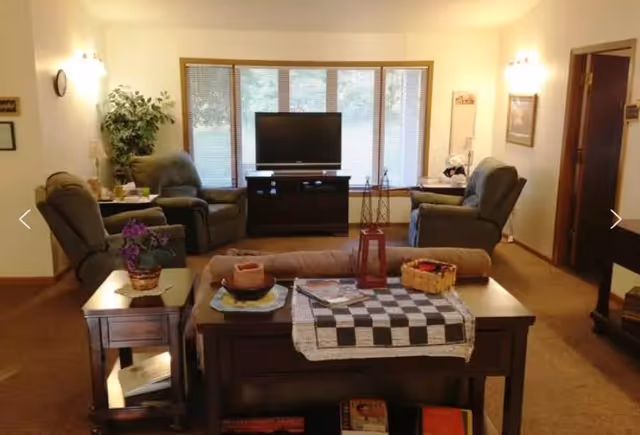 A cozy living room with four recliner chairs arranged around a TV on a stand in front of a large window with blinds. There is a wooden table with a checkered cloth, a basket, and decorative items in the foreground, and a side table with a potted plant and lamp to the left. The room has beige walls and carpeted floor.