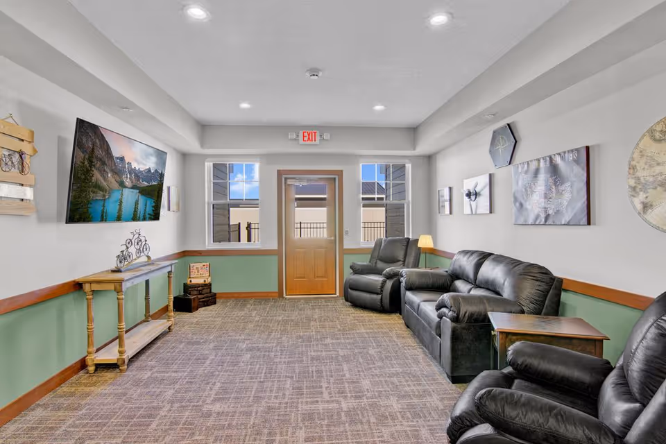 A cozy senior living common area with three black leather recliners and a matching sofa arranged along the right wall. The room has a carpeted floor, light gray walls with green wainscoting, and wooden trim. A wooden door with windows on either side is at the far end, letting in natural light. On the left wall, a flat-screen TV displays a scenic mountain and lake view, and below it is a wooden console table with decorative items. Various framed pictures and wall decor are hung on the walls.
