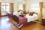A bright and clean bedroom in a care facility with two hospital-style beds, each with white linens and purple throws. The room has wooden flooring, a large window with beige curtains letting in natural light, and a small table with flowers and framed photos between the beds.