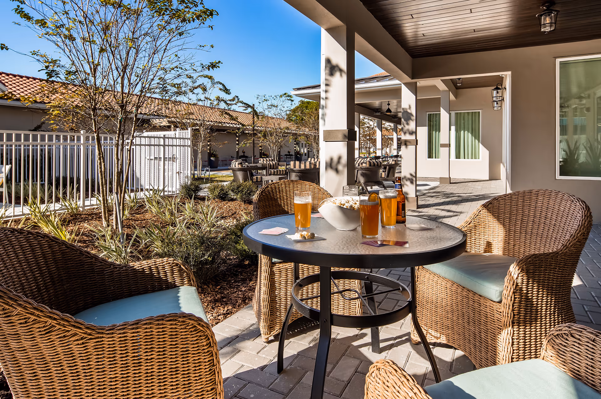 Outdoor patio area with wicker chairs and a round glass table holding four glasses of beer, a bottle, a bowl of popcorn, and playing cards. The patio is shaded by a roof and surrounded by plants and trees with a clear blue sky in the background.