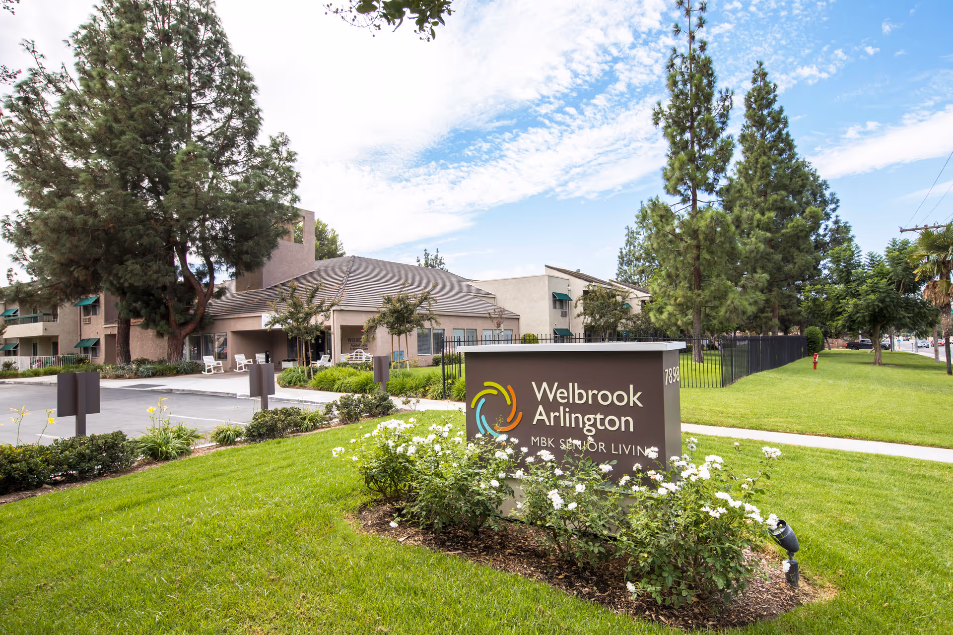 Entrance sign and front of a senior living community building with landscaped lawn and trees.