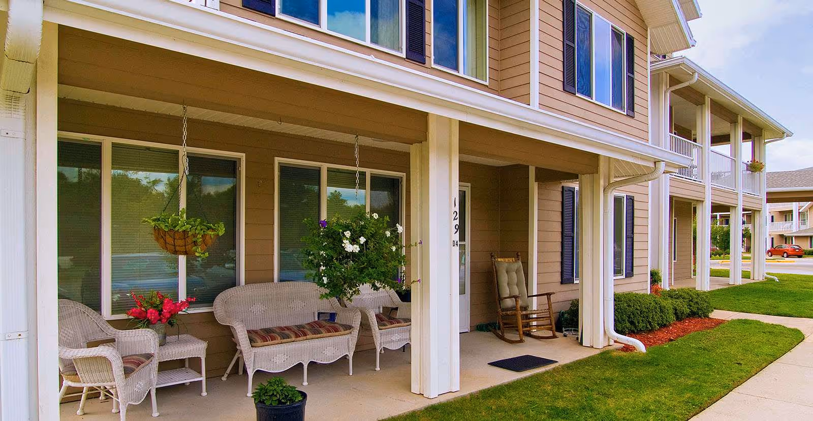 Covered front porch of a two-story beige senior living building with wicker seating, hanging flower baskets, a rocking chair, and a sidewalk.