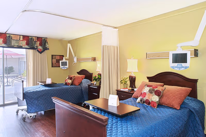 A bright and clean shared bedroom in a healthcare facility with two single beds covered in blue quilts and decorated with colorful pillows. Each bed has a bedside table with a lamp and a small plant. There are medical monitors mounted on the wall above each bed. A privacy curtain hangs between the beds. A sliding glass door leads to an outdoor patio area with tables and umbrellas.