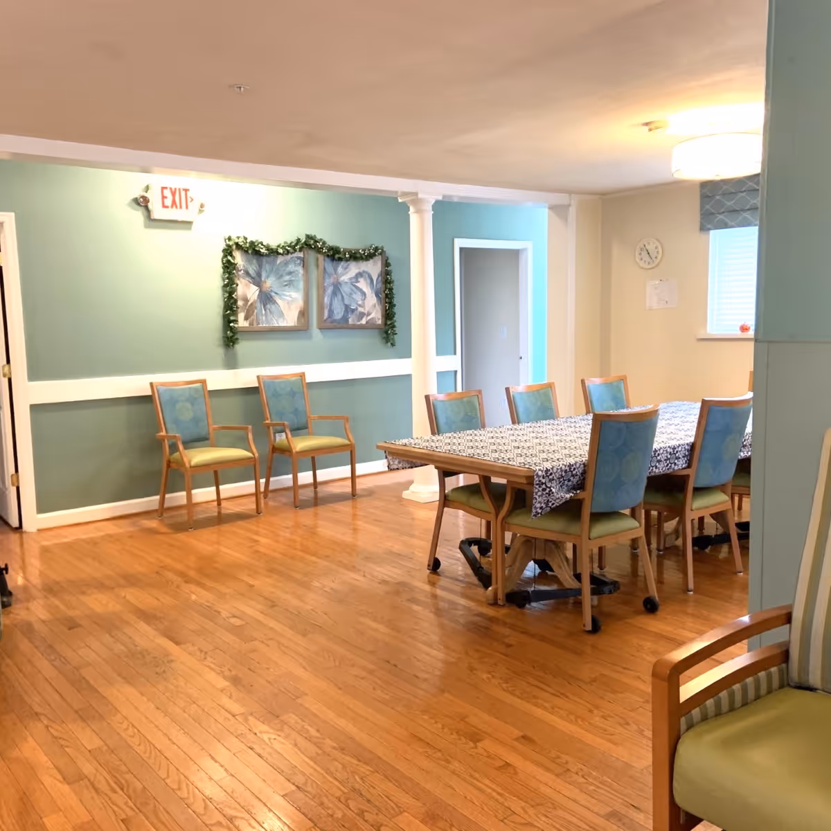 Dining area with a long table covered by a patterned tablecloth surrounded by blue-green chairs and hardwood floors.