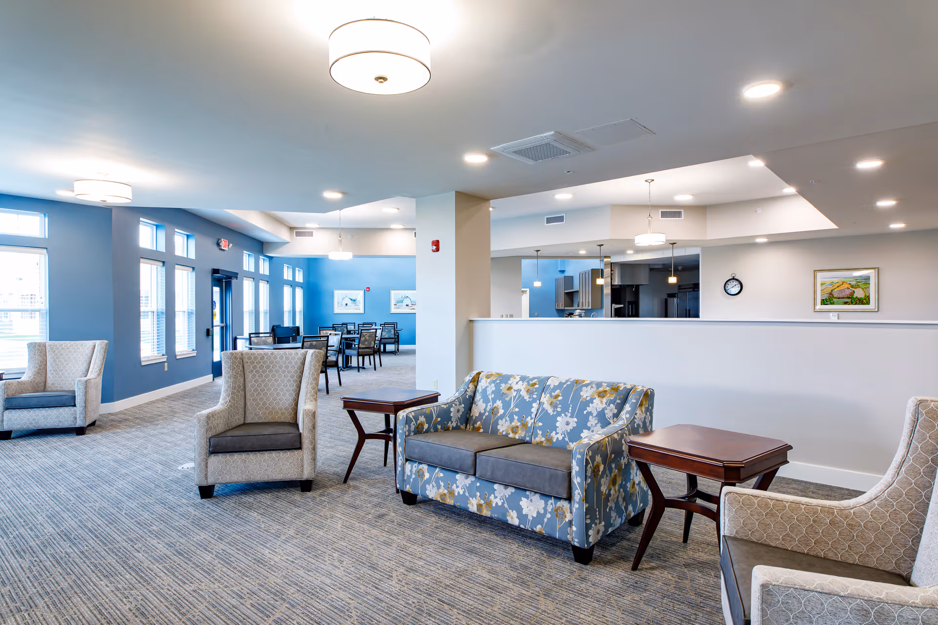 Well-lit senior living facility common area with a patterned sofa and armchairs, side tables, and a dining area visible in the background.
