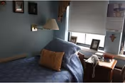 A small, cozy bedroom with a single bed covered in a blue patterned bedspread and a brown pillow. There is a nightstand next to the bed with a white telephone, framed photos, and other small items. A window with a white roller shade is above the nightstand, and the walls are painted light blue with a few framed pictures and a cross hanging on them.
