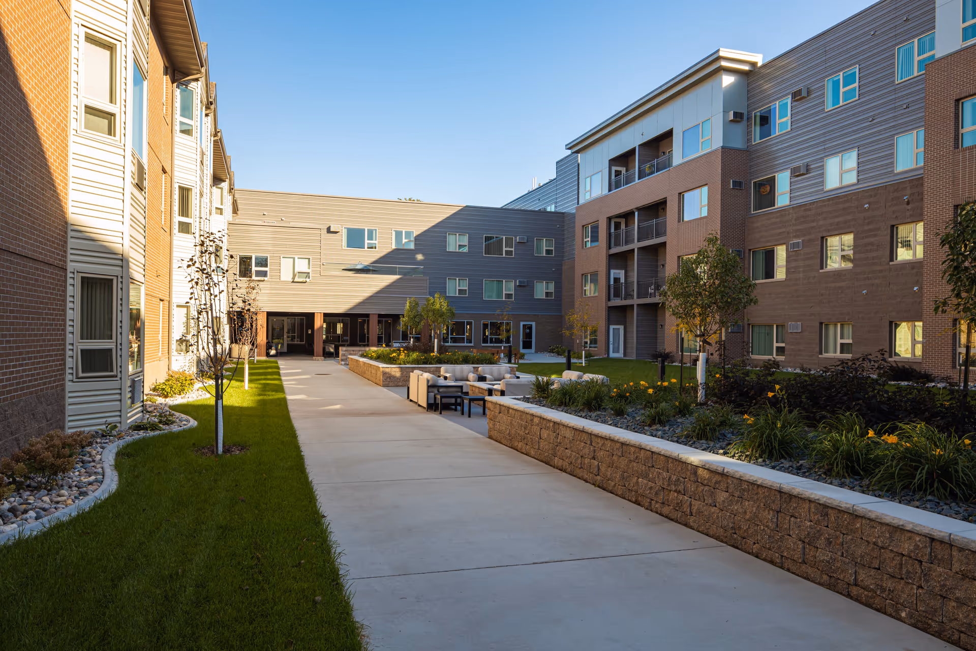 Outdoor courtyard area of The Linden senior living facility featuring a paved walkway, green grass, small trees, flower beds, and outdoor seating with cushioned chairs. The courtyard is surrounded by multi-story residential buildings with windows and balconies under a clear blue sky.