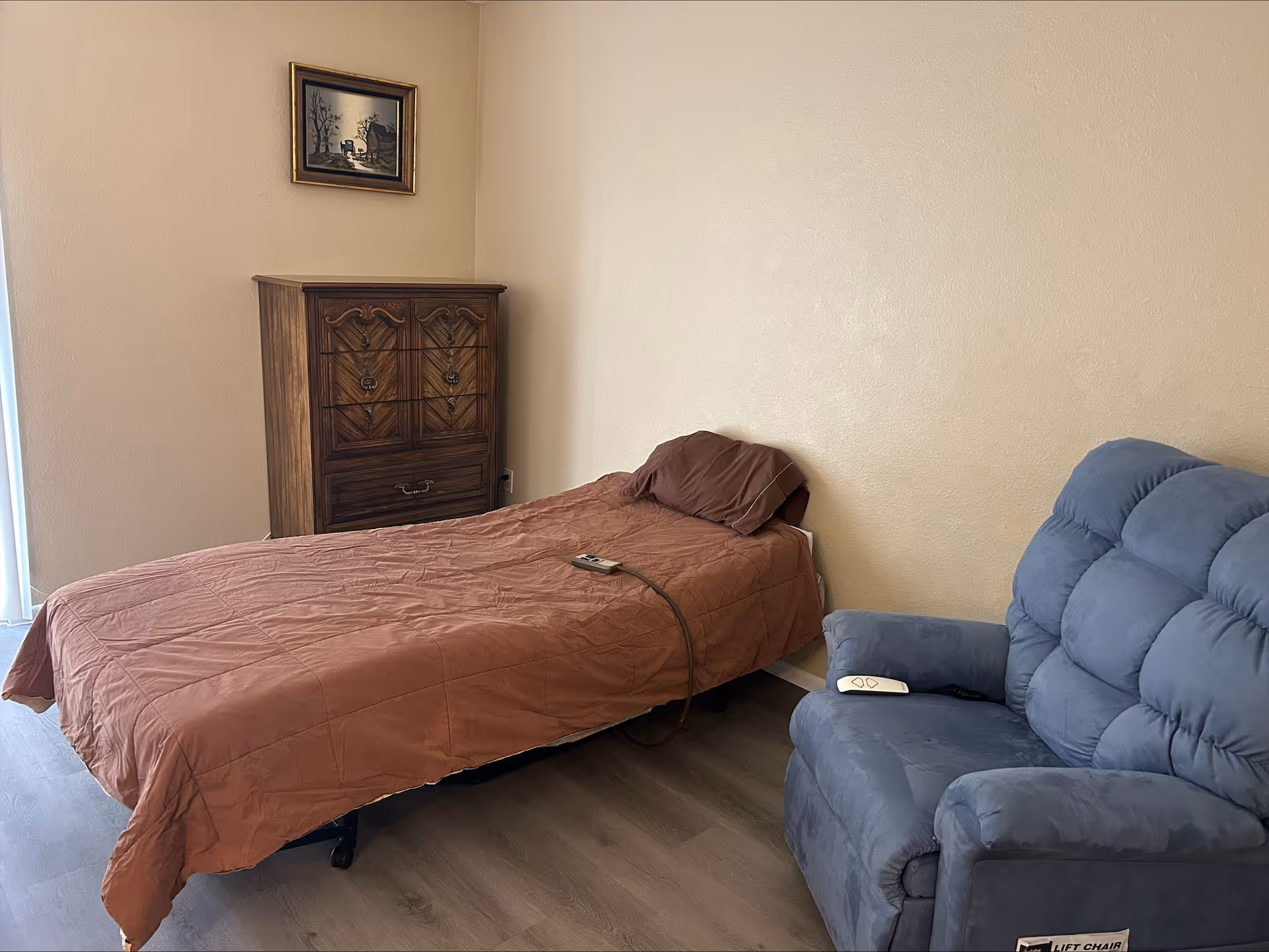Small bedroom with a single bed, wooden dresser, and a blue lift recliner against a beige wall.