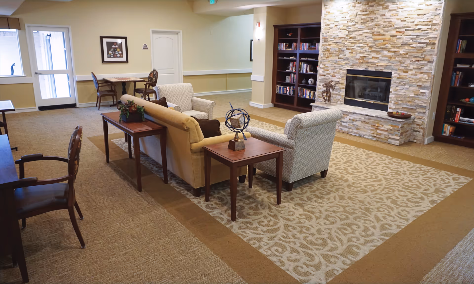 A cozy living room area in a senior living facility with a beige couch, two armchairs, wooden side tables, and decorative items. The room features a stone fireplace, built-in bookshelves filled with books, and a patterned area rug. There is a small table with two chairs near a door and a window in the background.