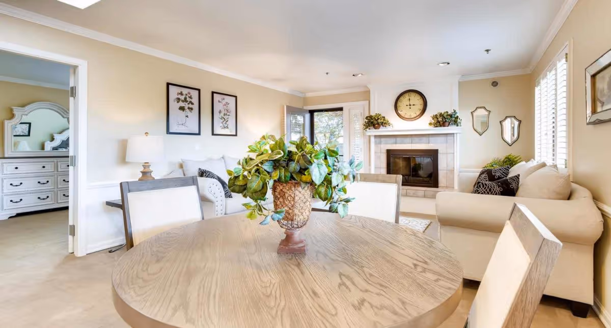 A cozy living room area with a round wooden table in the foreground holding a potted green plant. Behind the table are cushioned chairs and a beige sofa with black and white pillows. The room features a fireplace with a clock and plants on the mantel, two framed botanical prints on the wall, and a doorway leading to a bedroom with a white dresser and mirror.