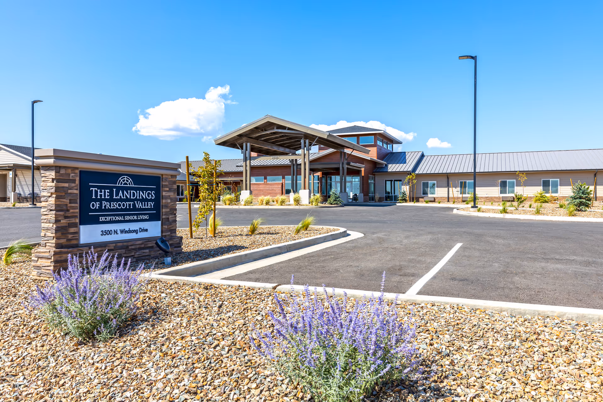 Front entrance and parking area of The Landings of Prescott Valley senior living facility with a sign, covered entryway, and landscaped gravel with purple flowers.