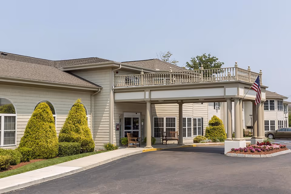 Exterior view of Benchmark Senior Living at Waltham Crossings showing the building entrance with a covered driveway, neatly trimmed bushes, an American flag, and a flower bed near the driveway.