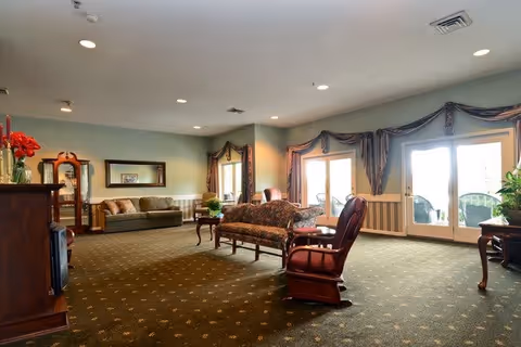 A spacious living room area with patterned carpet, multiple seating options including a floral sofa, a wooden armchair, and a beige couch. The room has large windows and glass doors with decorative curtains, allowing natural light to enter. There is a wooden grandfather clock, a mirror on the wall, and a wooden cabinet with a vase of red flowers on top.