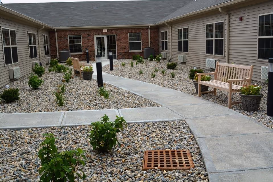 A curved paved walkway runs through a gravel courtyard planted with small shrubs and benches, framed by the single-story Aster Place building.