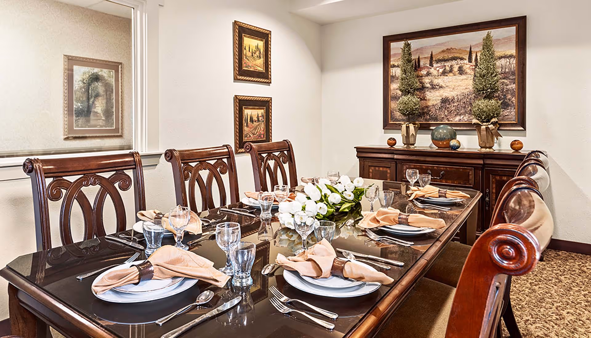 A formal dining room with a dark wooden table set for six people, featuring beige napkins, glassware, and white plates. The table has a centerpiece of white flowers. The room has wooden chairs with intricate backs, a sideboard with decorative items including two small topiary plants and a globe, and framed landscape paintings on the walls.